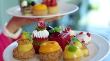 Human hands hold two plates of dessert cakes for two persons. All cakes with different flavors and interesting shape. There are raspberries, peach, pineapple, lemon, cherry.