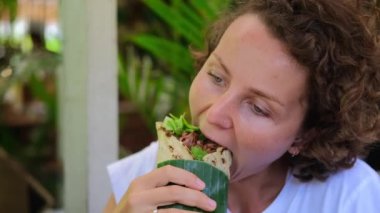 A young woman is sitting in a street cafe. In her hand she holds a flatbread stuffed with meat and vegetables inside. The roll is wrapped in a palm leaf. The woman takes a bite.