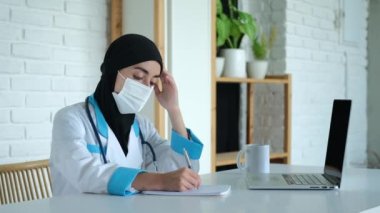 Siyah çarşaflı yorgun bir doktor ve işyerinde maske takan bir doktor laptopun olduğu bir masada oturuyor, doktor klinikteki hastaların kayıtlarını dolduruyor. Müslüman hastanesinde çalışıyorum..