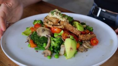 On a white plate, top view, there is a portion of fresh salad of vegetables, mushrooms, meat. A woman moves her fork in her plate, choosing what to eat first. Healthy and delicious food