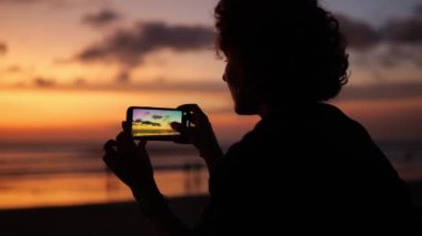 Silhouette of a woman blogger from the back against the backdrop of a sunset. A woman holds a smartphone with both hands and shoots the onset of twilight.