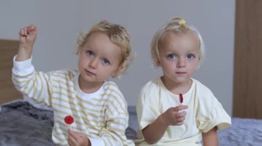 Two sisters sit facing the camera. Both babies are sitting on the bed, each of them has a lollipop in their hand. From time to time they lick them. Two very active children.