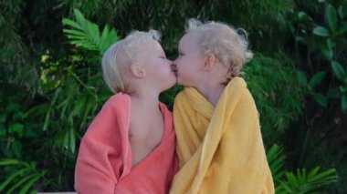 Little twin girls are sitting in bright towels on a bench after swimming in the pool. They are happy and cheerful, look at each other, laugh and kiss.