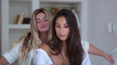 Two beautiful girlfriends are sitting together on the bed in the bedroom, the blonde hugs the brunette from the back. Two happy girlfriends spend weekend together in a room.