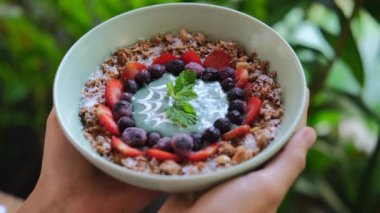 Delicious breakfast in the form of muesli with berries is a healthy breakfast that will fill your day with energy and vitamins. In female hands, on a green background, they hold a plate of porridge.