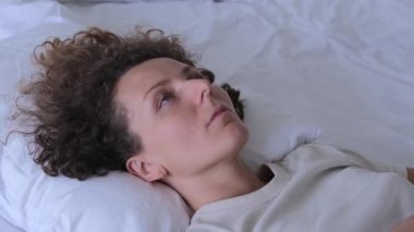 Sad woman lies on the bed with depression and migraine. A woman puts her palms to her head, overcoming pain and despair. Tired woman lies on the bed on the pillows in the bedroom.