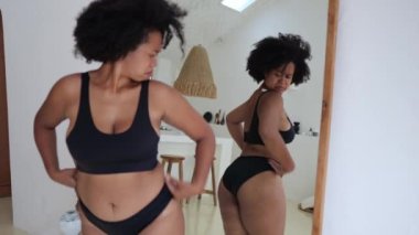 An overweight black woman looks at herself in the mirror and at her fatty parts of the body, the woman has stress due to being overweight. African American woman with obesity.