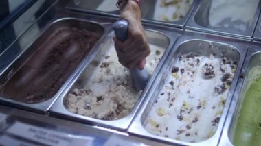 A showcase with different ice cream in containers, a woman scoops ice cream into a waffle cup with a spoon. Consumption of sweets with a high content of sugar and milk fat.
