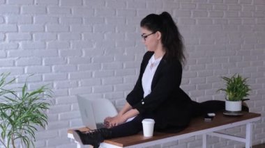 An employee of the office, with a business interior, may well boast of a stretching twine exercise, sitting on a table and typing in a laptop, looking at the documents sipping from a glass.