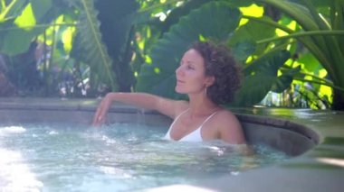 Beautiful woman with curly hair takes a jacuzzi in a spa hotel on vacation. A woman relaxes in a bath surrounded by lush tropical foliage. Spa treatments on vacation to restore strength.