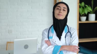 A friendly smiling female doctor in her office is waiting for new patients. A Muslim woman from the Middle East in a black hijab and medical coat works as a doctor in a private clinic.