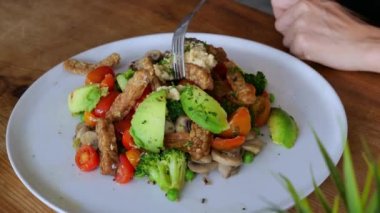 On a white plate, top view, there is a portion of fresh salad of vegetables, mushrooms, meat. A woman moves her fork in her plate, choosing what to eat first. Healthy and delicious food