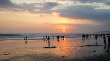 In the background of the ocean at sunset in the rays of the sun leaving the horizon, a lot of people are walking along the beach. There are many clouds in the sky
