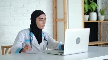 Tesettürlü bir doktor ve hastane üniformalı bir doktor hastayla video bağlantısı yoluyla iletişim kuran bir laptop kamerasına bakar ve belirtileri kaydeder. Hastanın çevrimiçi danışma.