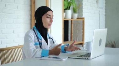 A female doctor in a hijab is seeing patients online using a webcam. A Muslim female doctor consults a patient via the Internet. A clinic that accepts clients of different faiths. 