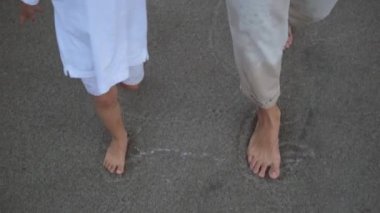 In slow motion, two pairs of bare, tanned feet for women and children walk on the wet sand on the beach. Their clothes are wet from walking near the water.