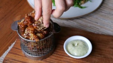 Close-up top view of a mans hand with two fingers, takes a small piece of meat cooked in teriyaki sauce with sesame seeds and dips it in a thick white sauce with pieces of greens