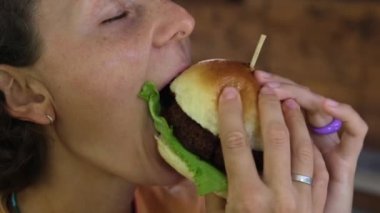 A young woman bites a delicious hamburger with appetite. The concept of unhealthy eating with the consumption of high-colored food that leads to obesity and health problems.