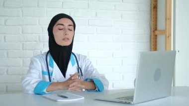 Work in a Muslim hospital. A tired doctor in a black veil and a medical gown at the workplace sits at a table on which there is a laptop, a woman has a headache from overexertion.