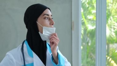 A tired Muslim nurse in a black hijab takes off her protective mask and sighs heavily after a work shift in the infectious ward with those infected with the coronovirus. Muslim doctor woman.