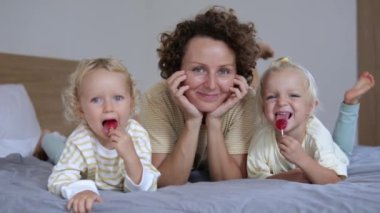 A young mother with her two twin daughters lie on the bed and look at the camera. The girls have one lollipop in their hands and they lick them with pleasure.