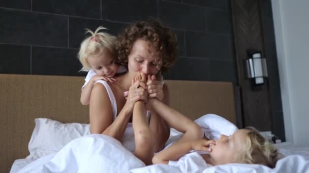 一位年轻的母亲坐在旅馆房间的床上 旁边是两个女孩的复制品 女人握住其中一个女孩的双腿 亲吻她的脚 — 图库视频影像