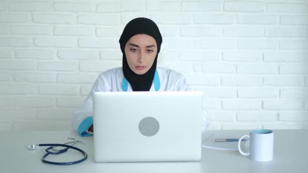 ヒジャーブのイスラム教徒の医師は 診療所を離れることなく患者のオンライン相談を行います 女性医師は自宅の患者を助けるためにインターネットとラップトップを使用しています — ストック動画