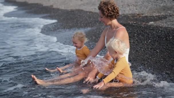 一位年轻的白人妇女带着两个小孩坐在海滩的边缘上 它们被温暖的海水冲刷着 一个女人教孩子们享受这些小事 — 图库视频影像