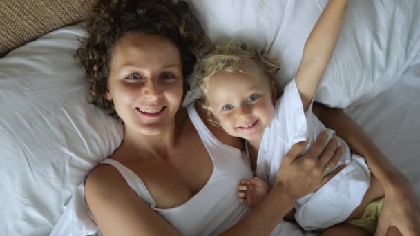 快乐的单身妈妈和小女儿躺在床上 躺在大枕头上 看着上方的相机 甜甜地微笑着 那女孩说了些什么快乐的家庭观念 — 图库视频影像