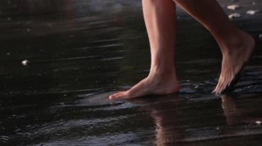 Naked female legs gently step on the wet sand and move towards the sea water. Another wave runs up to their ankles, giving them a pleasant coolness. Feet continue to walk already in the water.