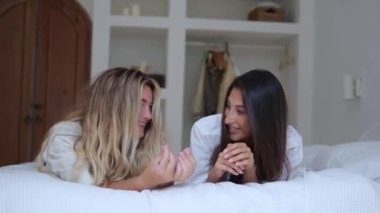 Two female friends lie on the bed together and laugh. A blonde with a brunette spend time together in the bedroom laughing to them having fun together. Beautiful girlfriends spend vacation together.