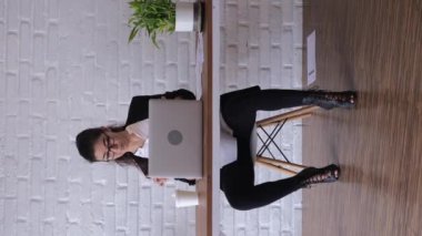 The employee has excellent inspiration to perform spontaneous dance movements, taking a sip of a drink, sitting at the table Crazy office plankton. vertical video. The woman is dancing in the office.