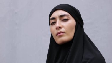 Close up of serious focused face of Armenian woman in black hijab on gray background. Face of feminist Islamic woman with black headscarf on head. Feminism in Islamic Countries, Muslim Feminist Woman
