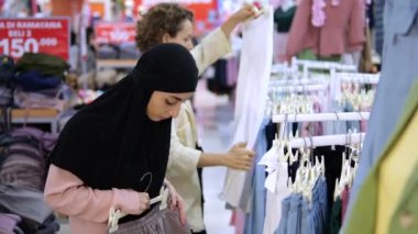 Alışveriş merkezindeki kadın giyim mağazasında pantolon ve pantolon seçen iki arkadaş. Kafkasyalı arkadaşıyla tesettüre girmiş bir Arap kadın hem kıyafet seçip hem de satın alır..