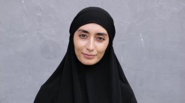 Siyah tesettürlü güzel bir Arap kadın gülümsüyor ve gri duvara doğru kameraya bakıyor. Yaklaş. Arap görünümlü bir kadının portresi büyüleyici bir gülümsemeyle tesettüre bürünmüş..