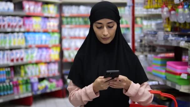 スーパーの真ん中で手にスマートフォンを持つヒジャーブのイスラム教徒の女性の肖像画 店内の女性は 購入についてのコンサルティング 電話でチャット 雑貨のある棚の近くの電話でイスラム教徒の女性 — ストック動画