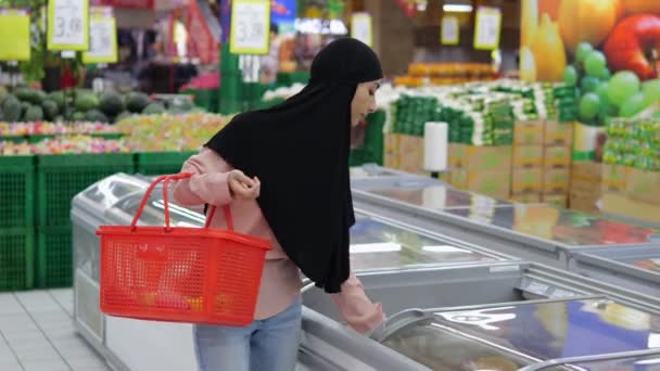 超市里的妇女从冰箱里取出冷冻产品 放在她的红色购物篮里 穆斯林妇女头戴头巾 头戴红色购物篮 在超市里买冷冻食品当晚餐 — 图库视频影像