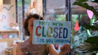 Kafkasyalı bir kadın dükkanın kapısının önündeki renkli kare tabelasını ters çeviriyor. Sabah, satıcı dükkanın kapılarını açar ve alıcıları davet eden camdaki tabelayı çevirir.