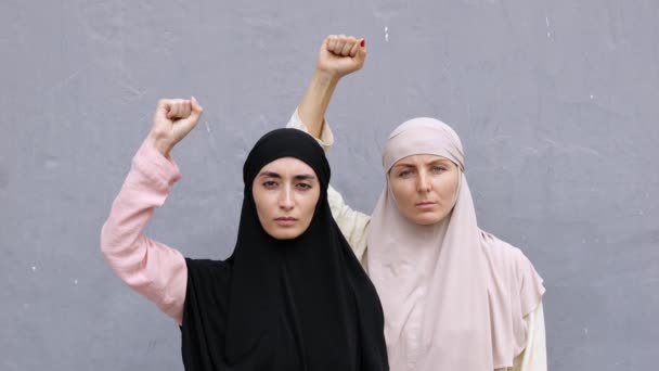 黒と白のヒジャーブを持つアラブ人と白人の女性が彼女の拳を上げて立っています 彼らの拳で立つ深刻で決定されたイスラム女性活動家の肖像画 抗議の象徴 — ストック動画