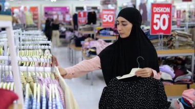 Khimar 'daki İslami kadın mağazadan bluz ve gömlek seçiyor. Müslüman kadın müşteriler bluzlarını bir mağazada karşılaştırarak neyi deneyeceklerini seçiyorlar. % 50 indirim, kadın giyim mağazasında eşya satışı..