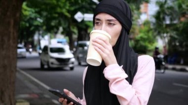 Siyah başörtülü Arap kadın caddede duruyor, kahve içiyor ve telefonunu kullanarak güzergah arıyor. Türbanlı İslamcı kadın yolunu bulmak için online haritalar kullanıyor. Başörtüsü olan kadın portresi