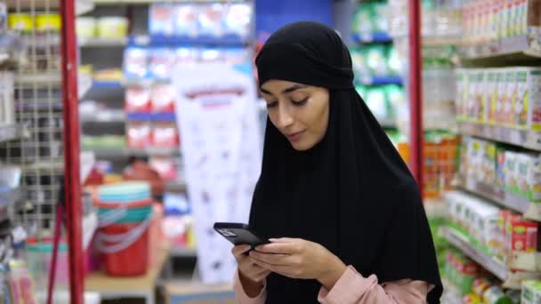 Красивая Женщина Хиджабе Посреди Полки Магазина Держит Руках Мобильный Телефон — стоковое видео