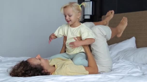 小さな女の子は楽しいダンスをしている母親の腹に座っています ママは娘を揺らすベッドの上に横になって足を振る ママは子供と遊ぶのが楽しい 母娘の余暇活動 — ストック動画