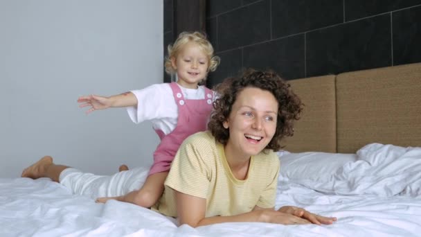 白人妇女和她的女婴躺在床上 笑着和笑着 可爱的女儿骑在妈妈的背上 亲切地搂着她的脖子 关心她 妈妈花时间和女儿在一起 — 图库视频影像