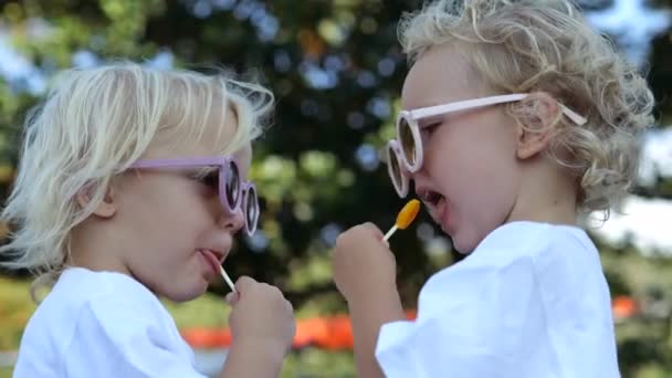 两个带眼镜的白人金发小孩的特写 孩子们在公园里吃焦糖棒棒糖 孩子们通过吃焦糖棒棒糖来争夺速度 儿童竞速游戏 — 图库视频影像