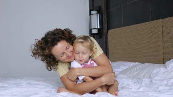 母は愛情を込めて娘を抱きかかえて頬にキスをし 思いやり 優しさを表現します 幸せな女性と女の子がベッドの上に座って 抱き合って 笑っています — ストック動画