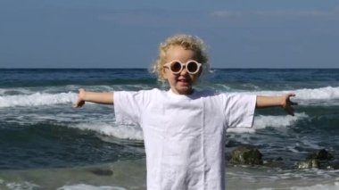 Kıvırcık saçlı, beyaz bir kız deniz dalgalarının kıyılarını yıkıyor. Moda kızı, kollarını açmış, gülümseyerek güneşlenir ve tatilin tadını çıkarır. Sıcak denizde hafta sonu.