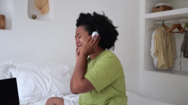 幸せな感情的な肯定的な黒人女性の笑顔 ヘッドフォンで音楽を聴く ヘッドフォンでお気に入りの音楽をお楽しみください ヘッドフォンで音楽を聴くベッドの上の多民族千年紀の女性 — ストック動画