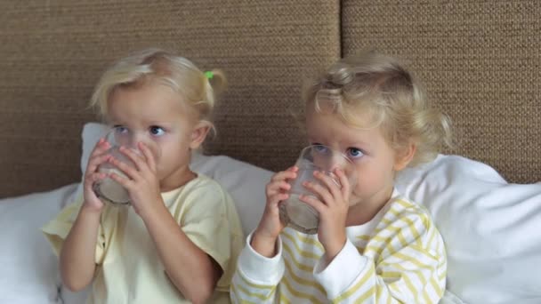 2人の白人ブロンドの双子の女の子は ベッドに座って 夏の暑さから渇きに対処するためにさわやかな冷たい健康的な飲み物を飲みます 双子の姉妹はガラスから飲むひどい渇きを取り除くクローズアップ — ストック動画