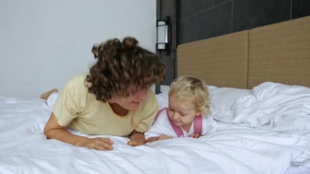 迷人的年轻卷发女子和可爱的金发小女孩白天躺在酒店的床上 母亲和女儿从酒店床上站起来看着摄像机的慢镜头 — 图库视频影像